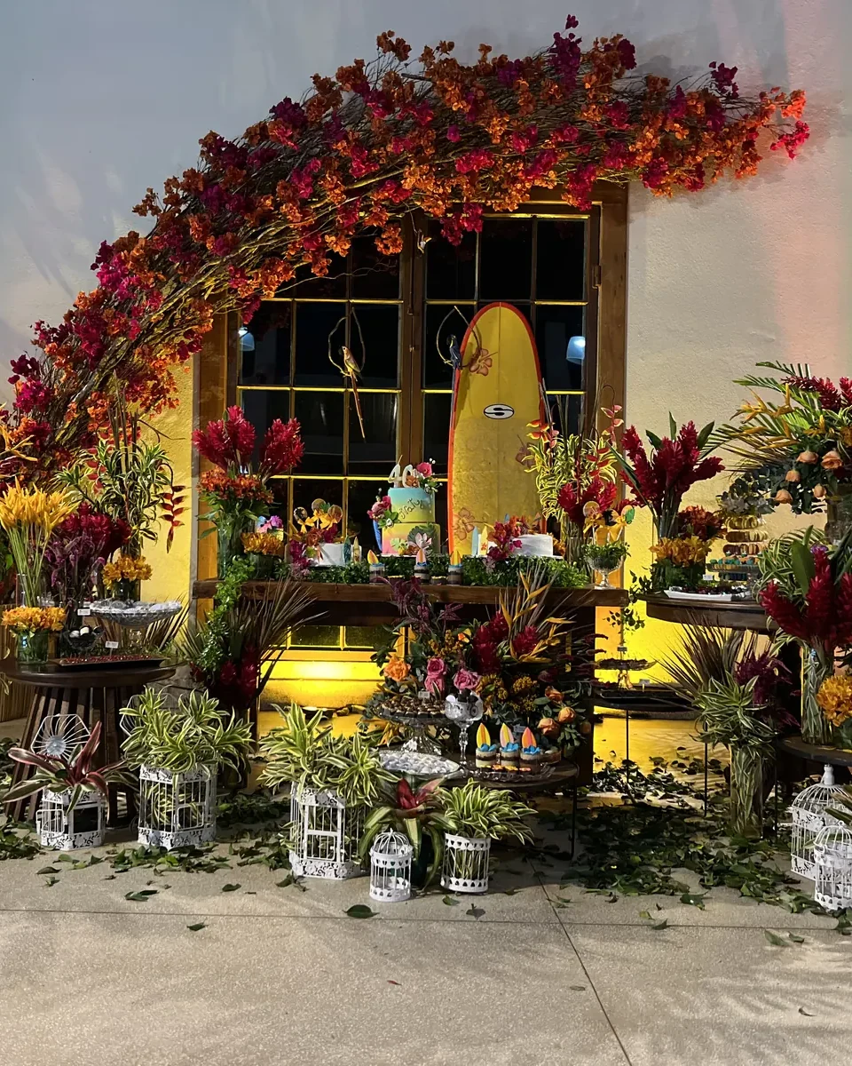 Mesa de doces do salão de festas montada sob um arco de flores, com arranjos tropicais em volta e a porta amarela iluminada ao fundo