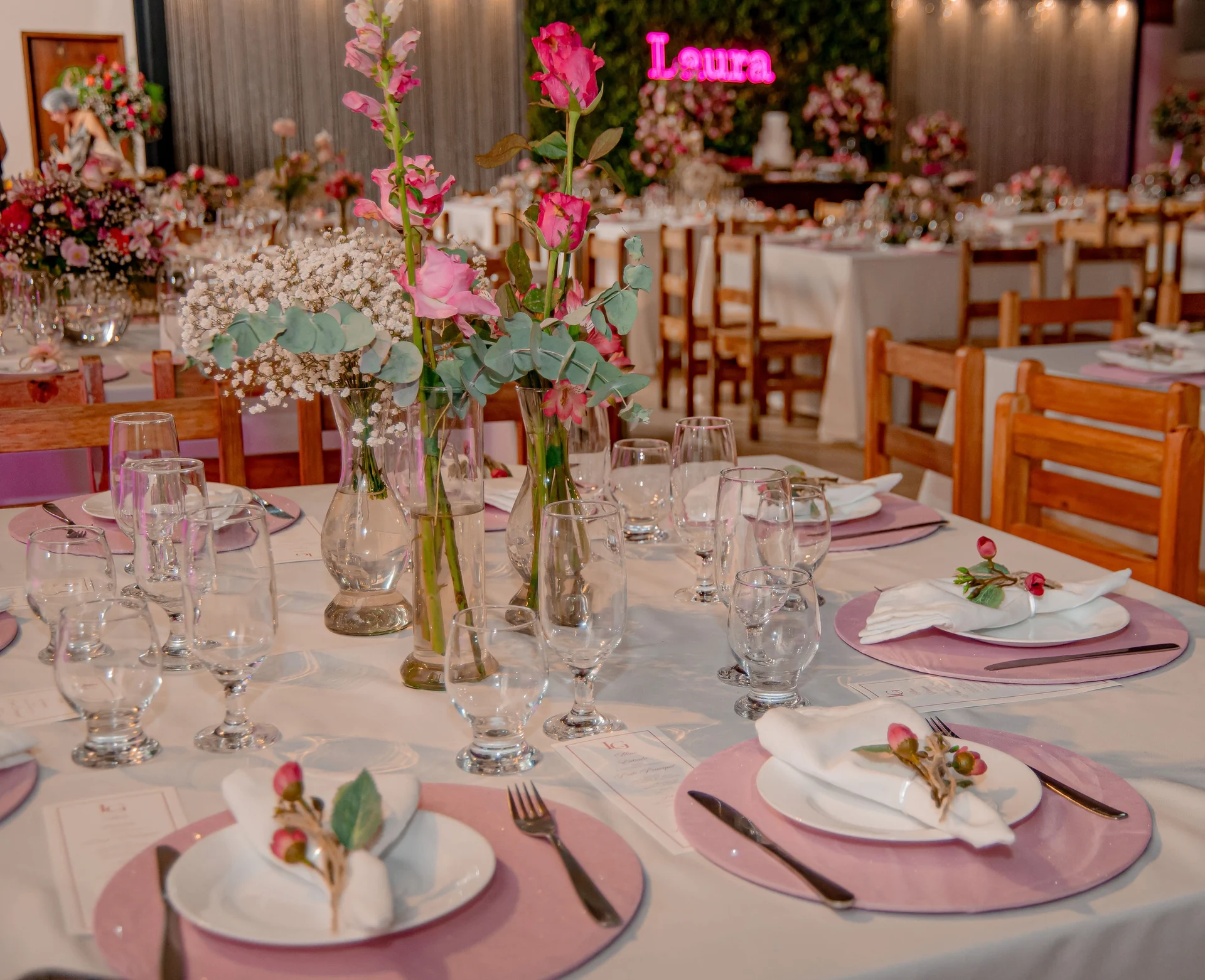 Mesas postas para uma festa de 15 anos no salão do Hotel Fazenda Minas Real, com sousplats rosa, arranjos de rosas e o painel de flores ao fundo
