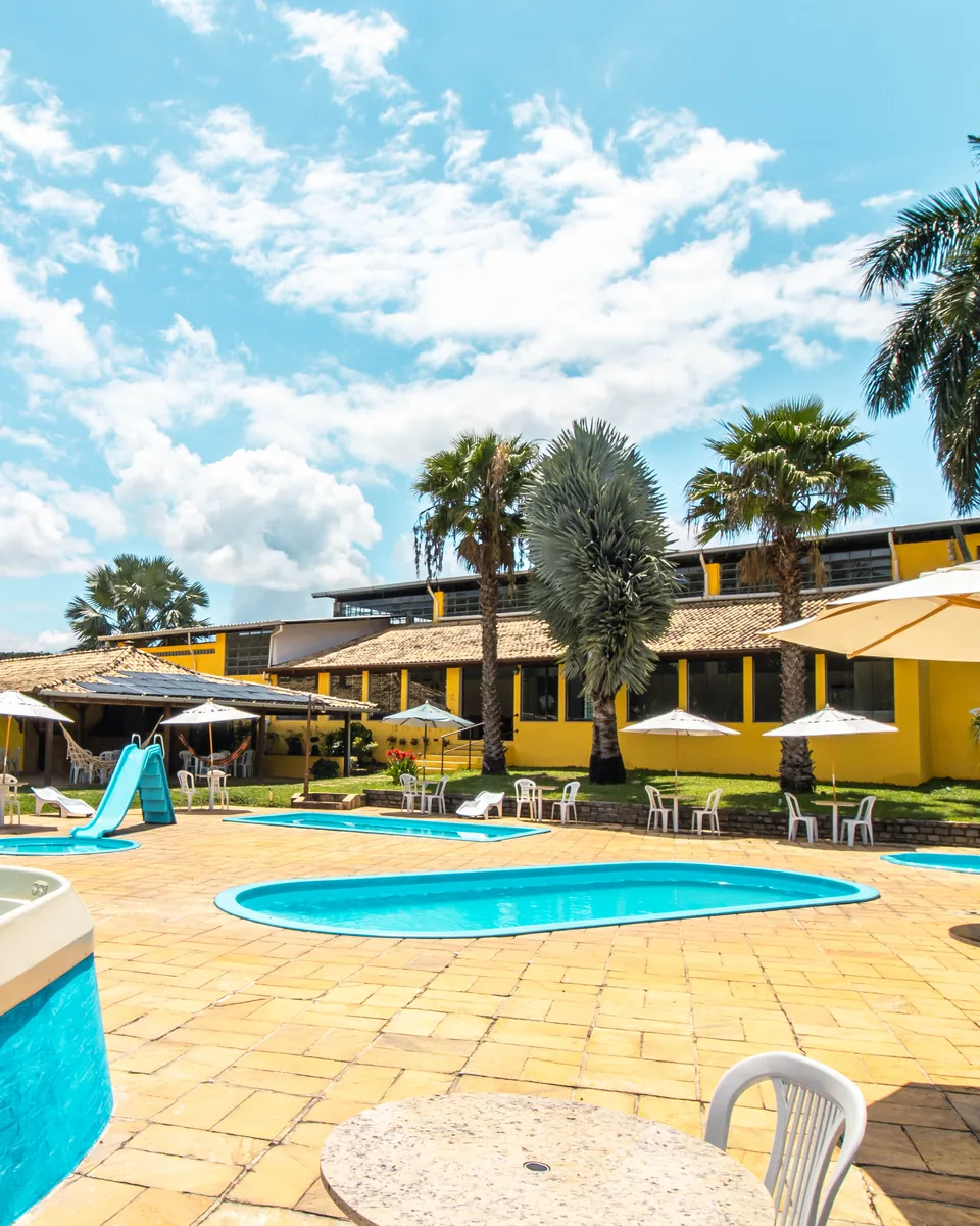 Piscinas e ofurô do Hotel Fazenda Minas Real, com guarda-sóis, palmeiras e o prédio amarelo do hotel ao fundo