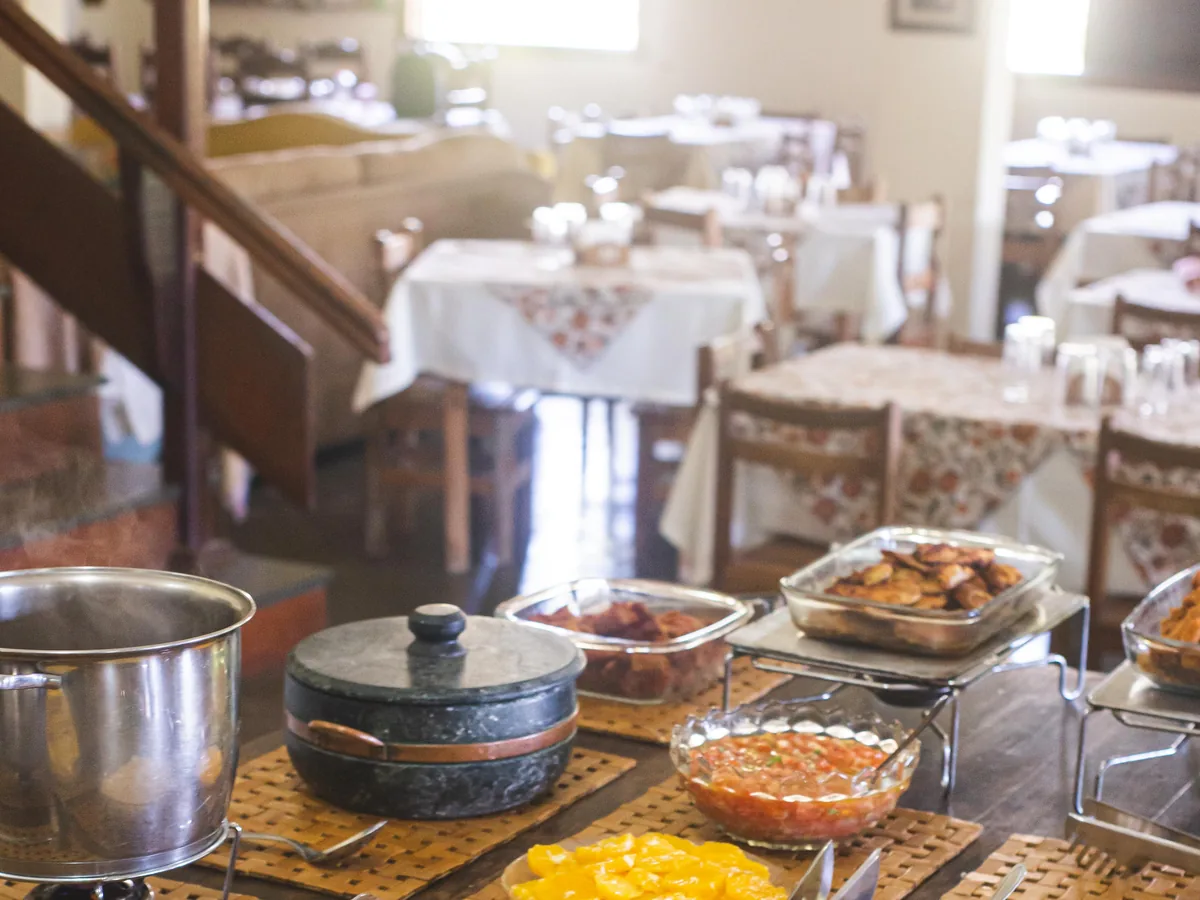 Mesa do buffet de almoço montada com angu, farofa, molhos e panelas, e o salão do restaurante posto ao fundo