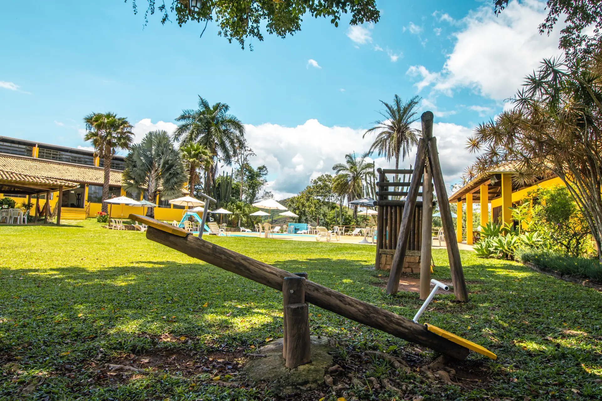 Gangorra e trepa-trepa no gramado do Hotel Fazenda Minas Real, com as piscinas, as palmeiras e a ala amarela ao fundo